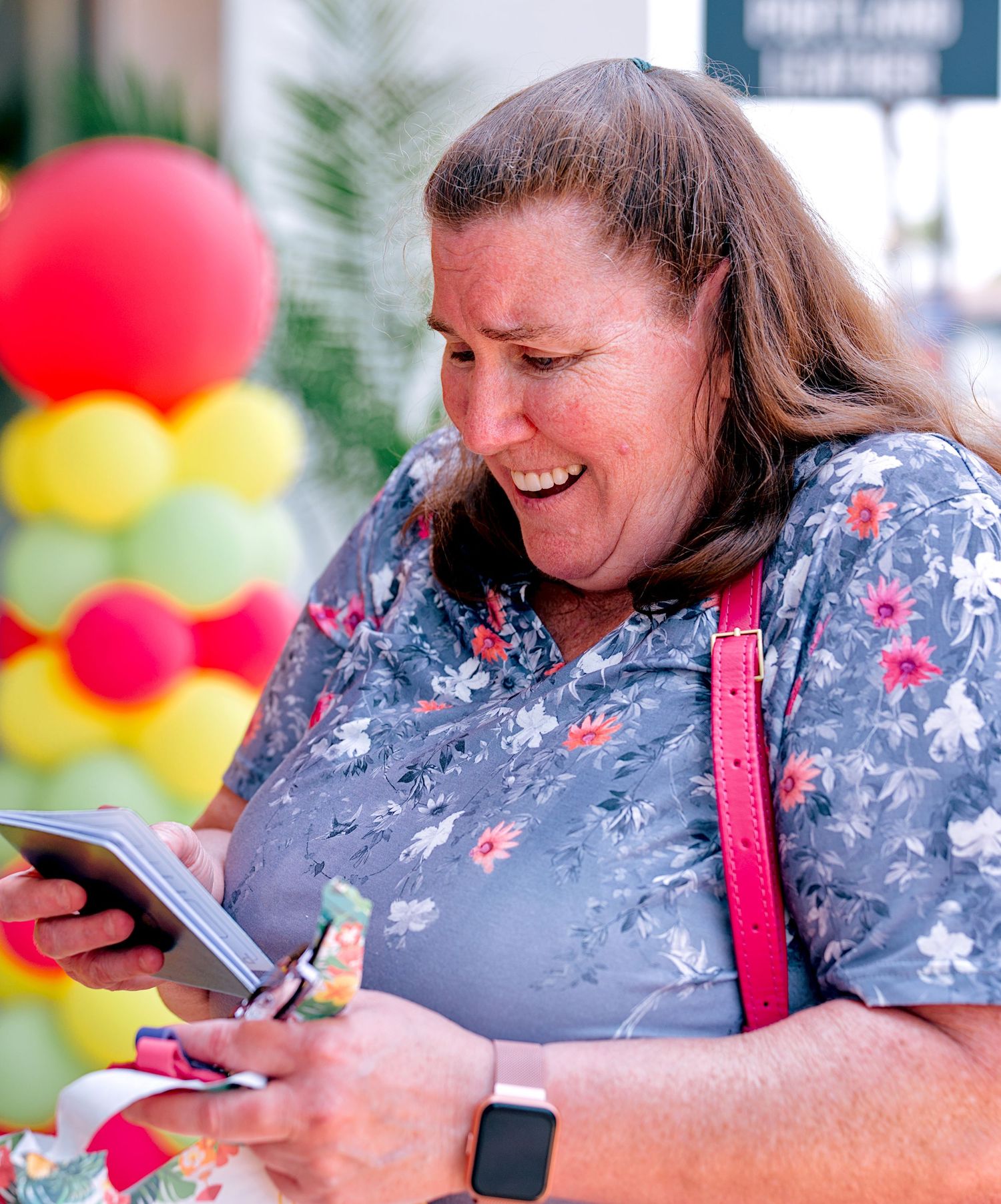A smiling woman in a floral blouse uses her phone outdoors near colorful balloon decorations.
