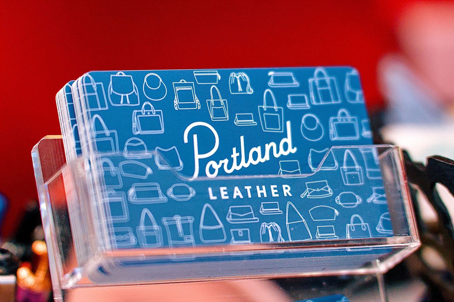 Portland Leather branded business cards displayed in a clear acrylic holder with blue design and bag illustrations.