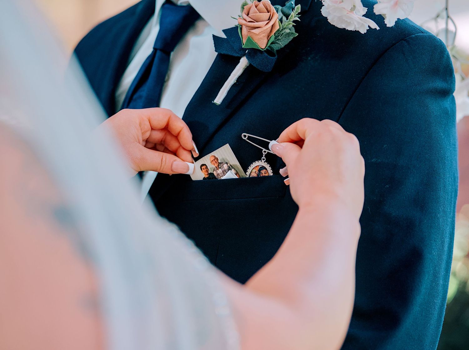 Groom in navy suit with floral boutonniere receiving a small photo keepsake in his pocket during wedding ceremony.