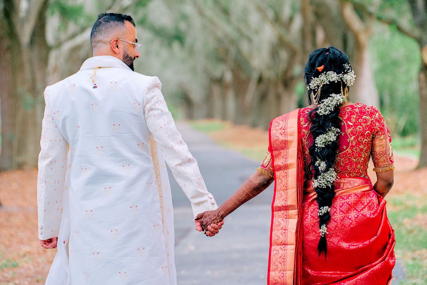 Indian bride and groom holding hands walking down tree-lined path in traditional wedding attire.