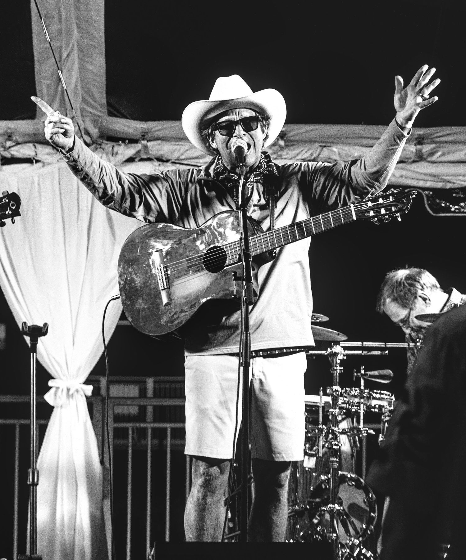A musician in a white cowboy hat performs on stage with an acoustic guitar at an outdoor concert.