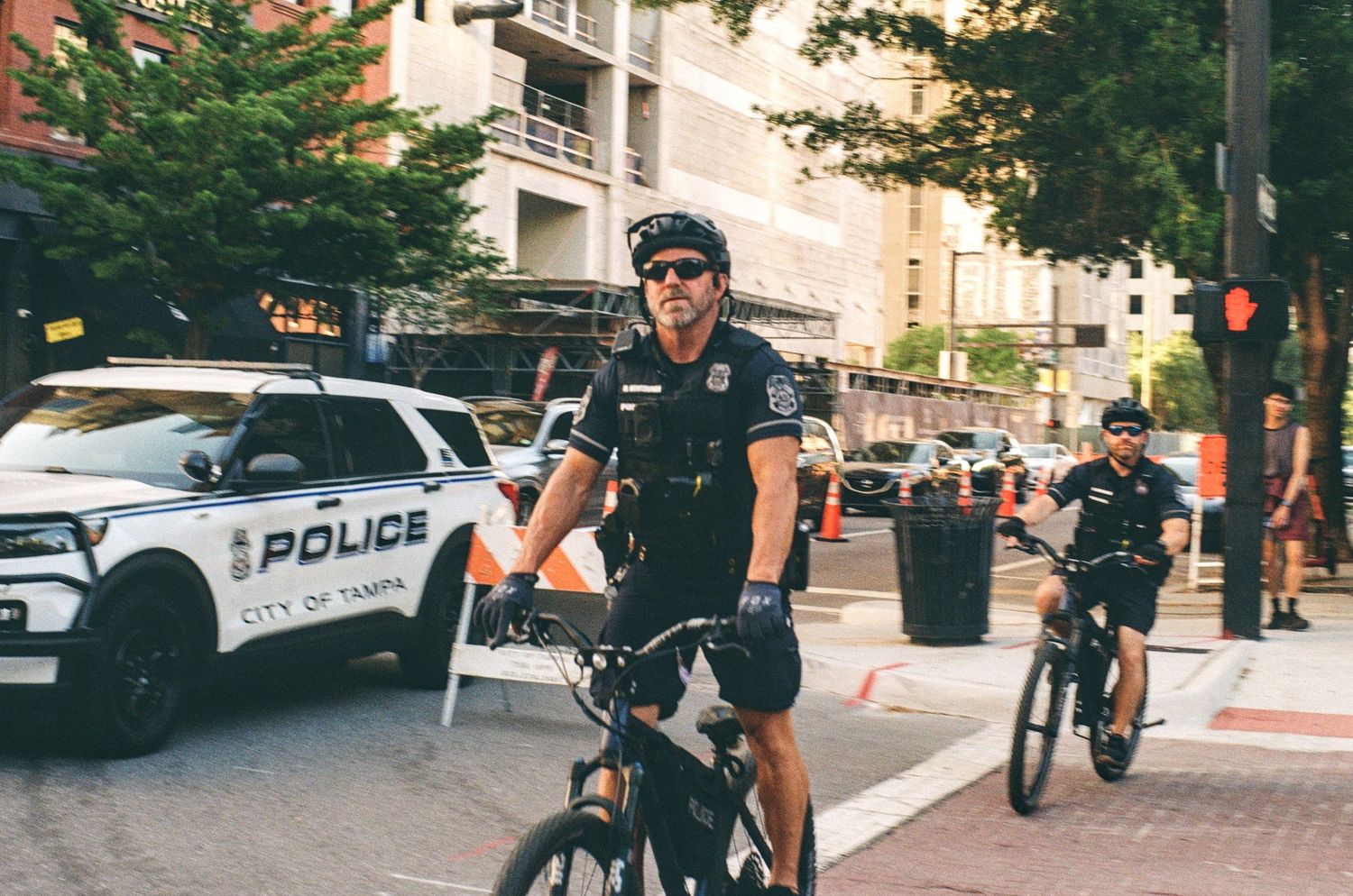 Two City of Tampa police officers patrol on bicycles near a police SUV on a busy urban street.