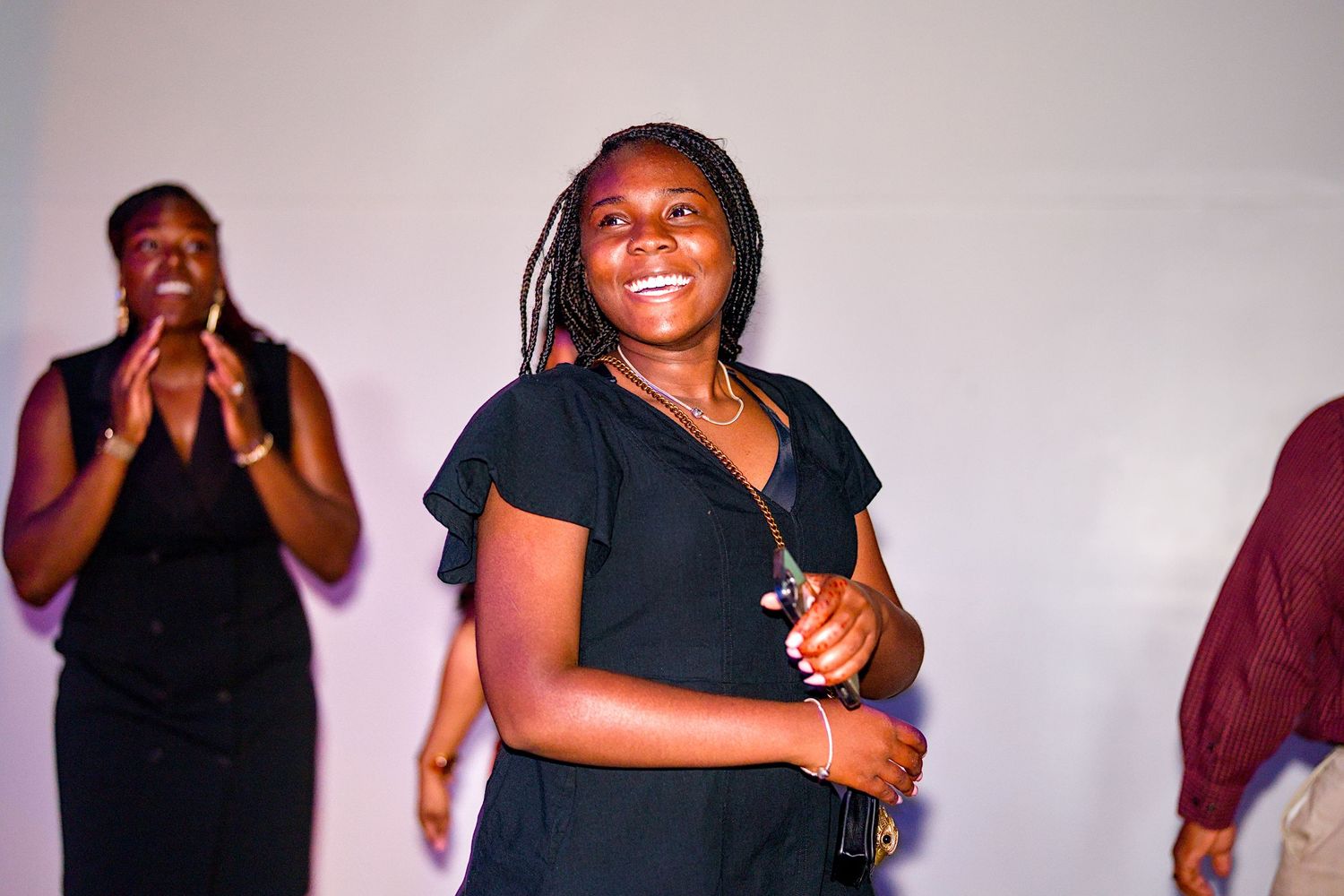 Smiling young woman in black dress dancing at an event with people clapping in the background.