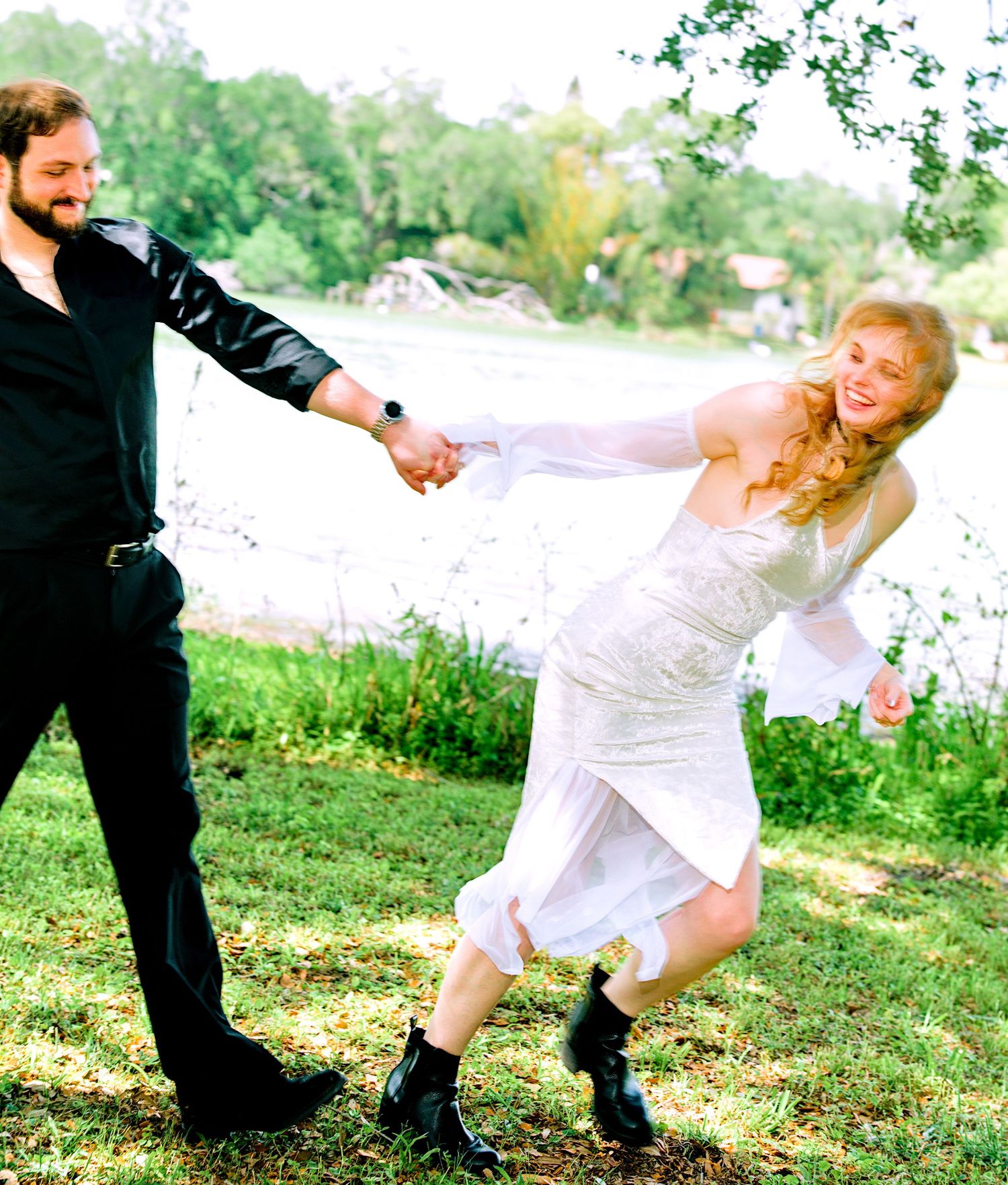 Joyful bride and groom dancing and spinning outdoors near a lake on their wedding day.