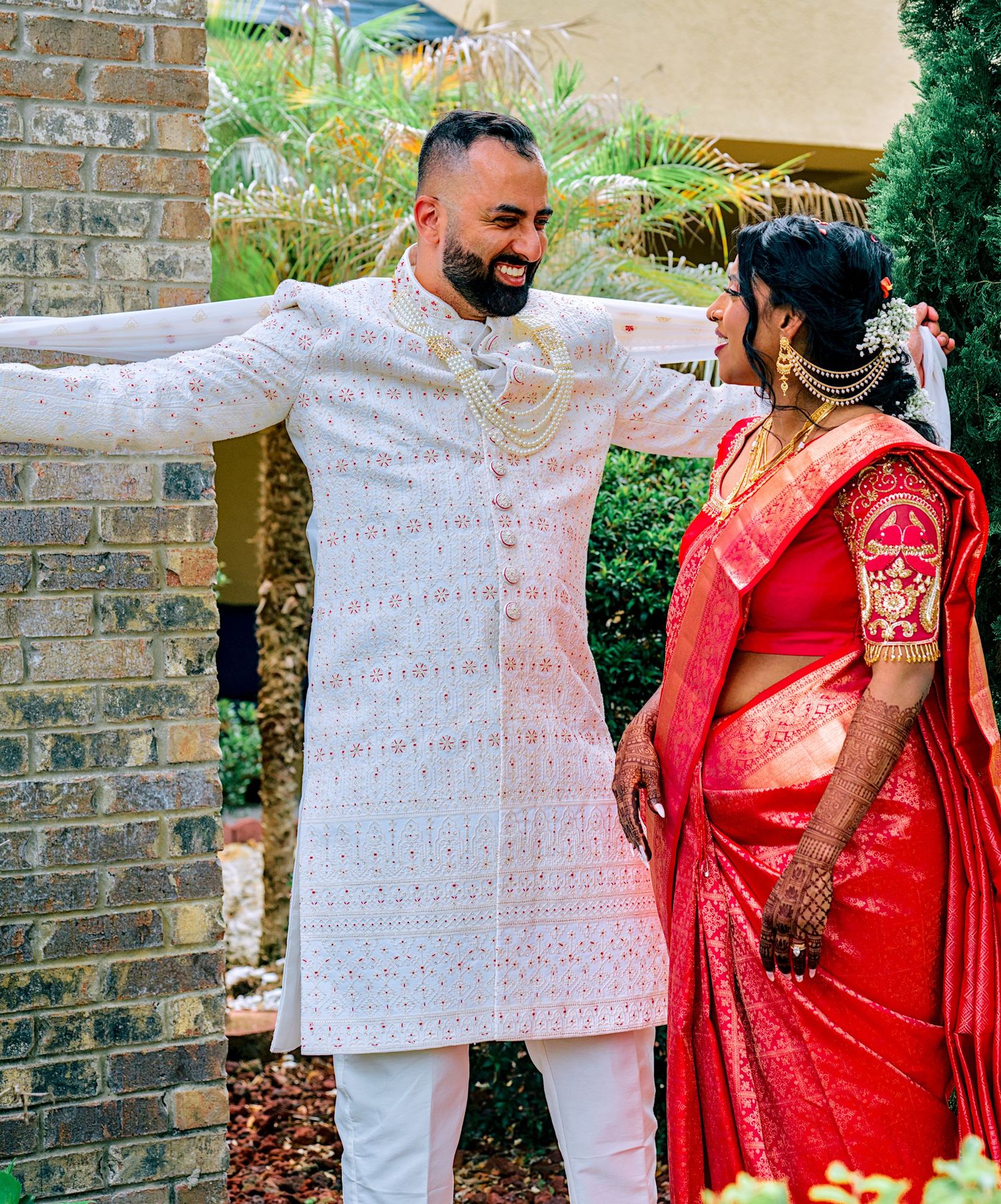 Indian groom in white sherwani and bride in red saree smiling together outdoors during traditional wedding ceremony.