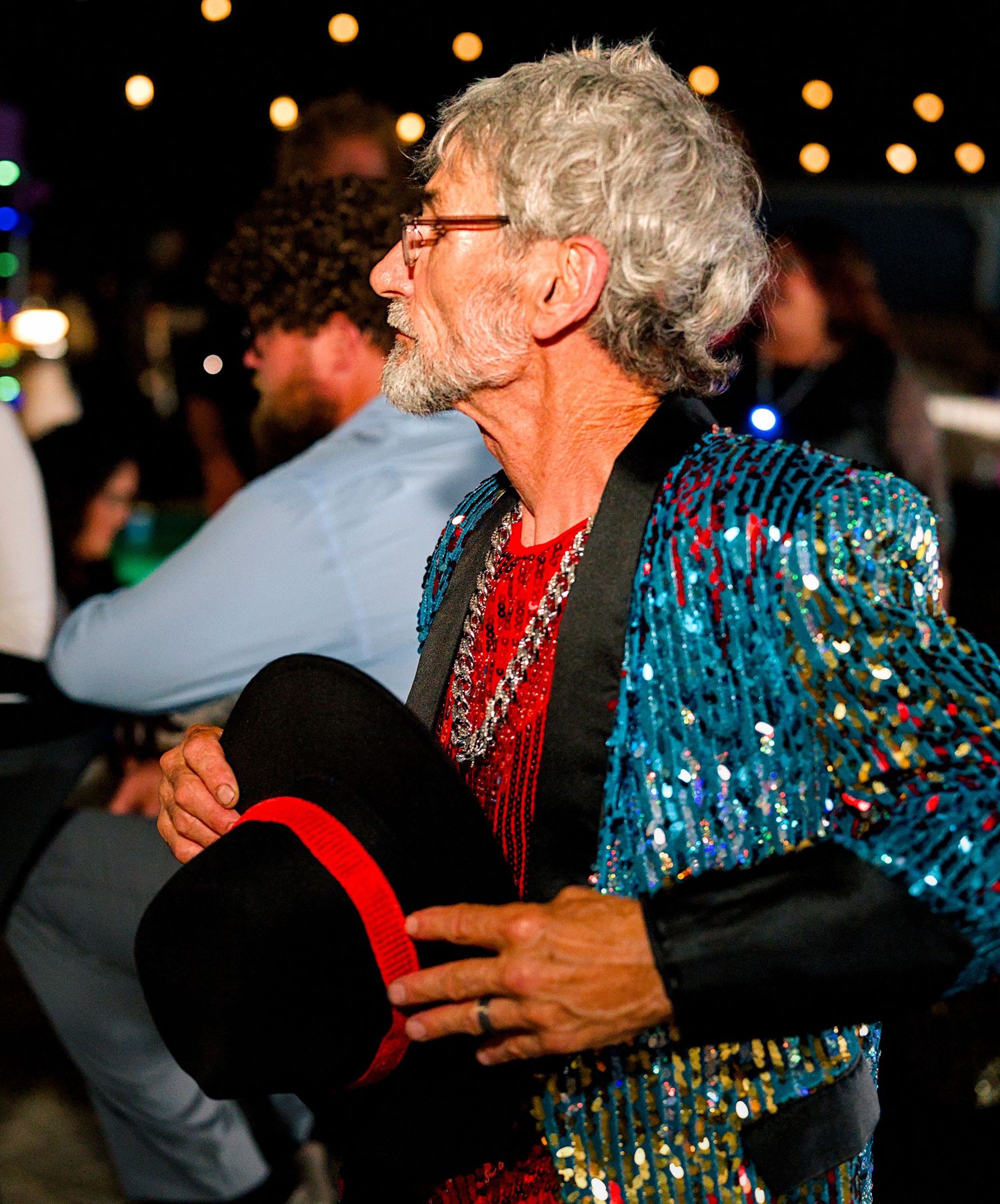 An older man in a sequined teal jacket holding a top hat at a festive outdoor disco-themed party at night.