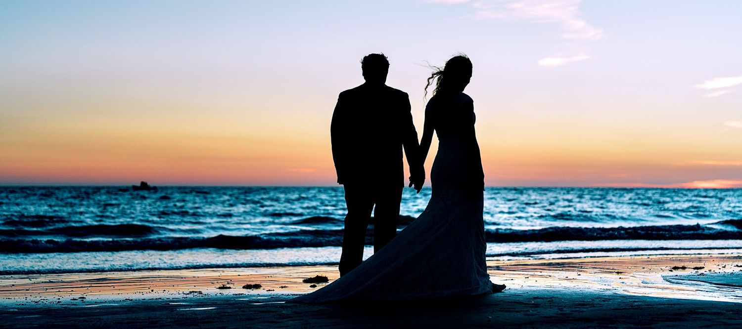 Silhouette of a bride and groom holding hands on a beach at sunset with ocean waves in the background.