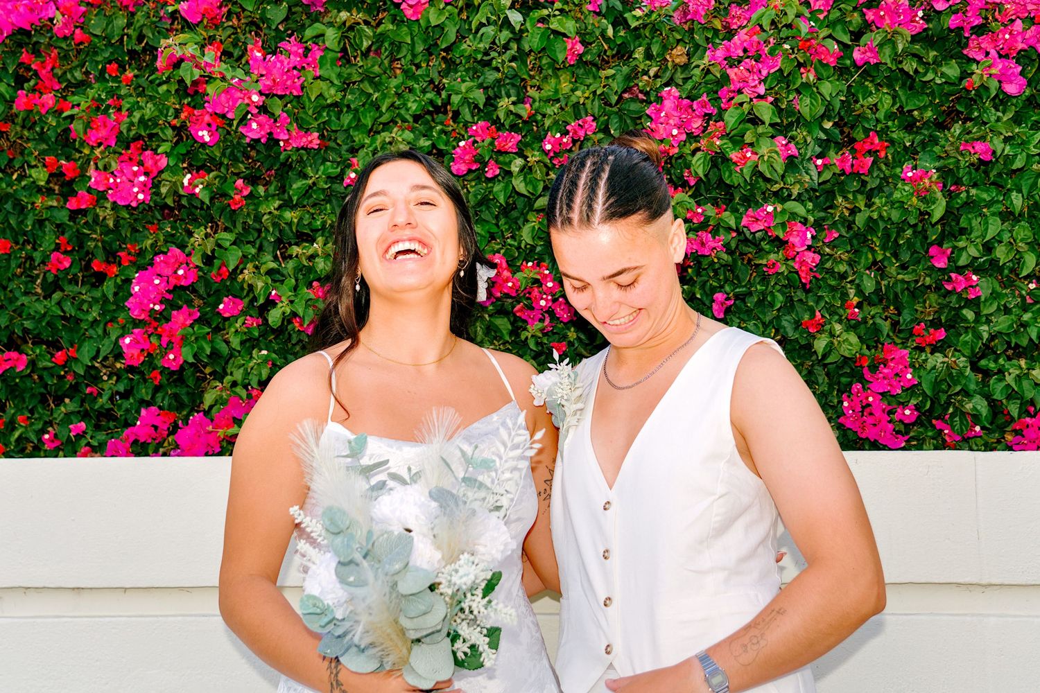 Two brides laughing and smiling together in front of vibrant pink bougainvillea flowers on their wedding day.