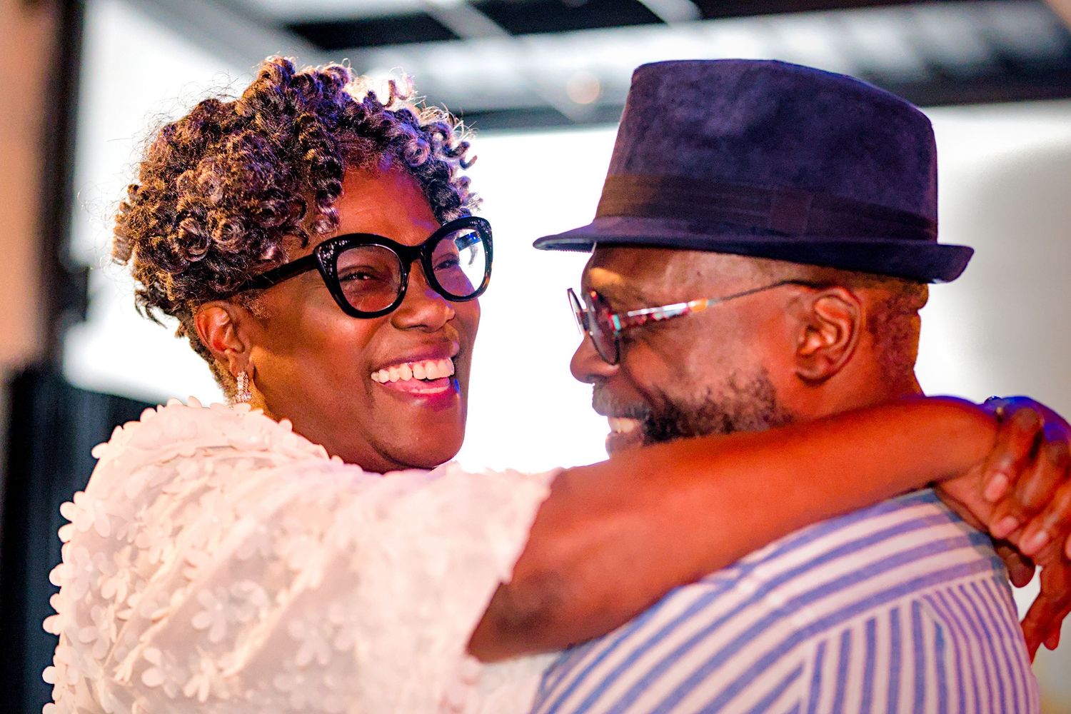 Smiling woman with curly hair and glasses embraces a bearded man in a blue hat at a joyful celebration.