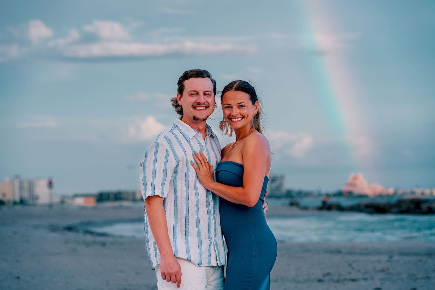 Happy couple embracing on a beach at sunset with a rainbow in the background.