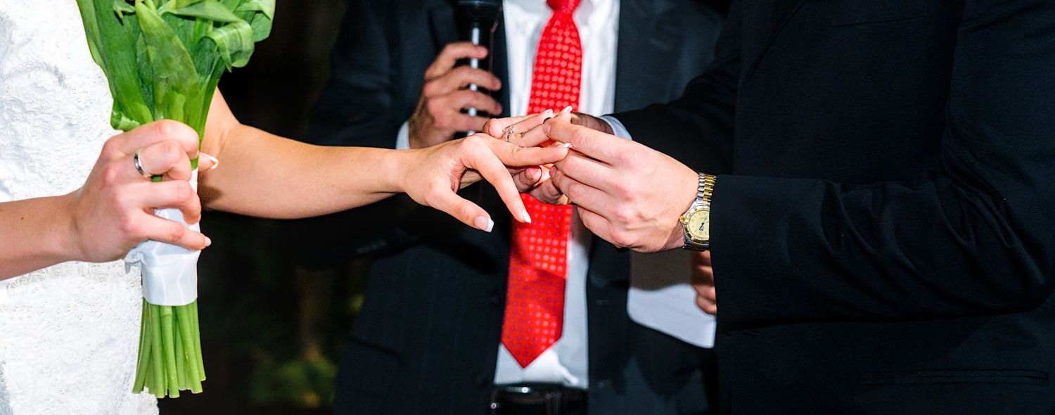Groom placing a wedding ring on bride's finger during ceremony, bride holding a white bouquet.