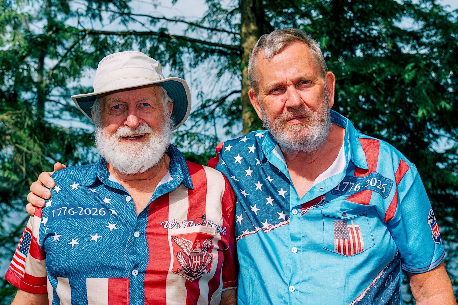Two men with white beards wearing patriotic American flag shirts pose together outdoors near green trees.