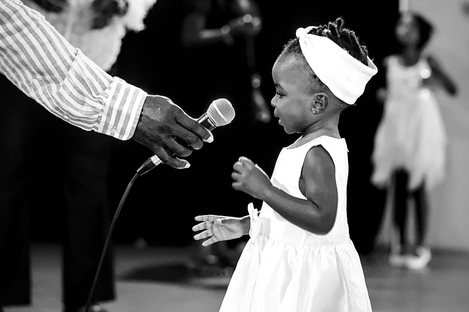 A young girl in a white dress reaches for a microphone at an event.