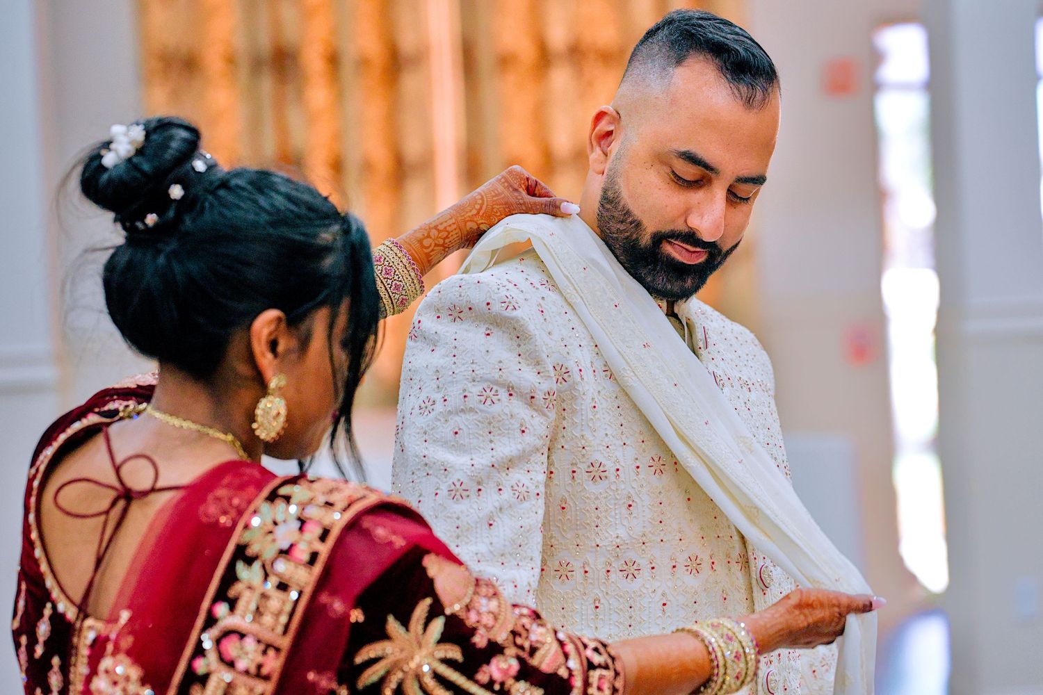 Woman in red lehenga draping white sherwani on groom during Indian wedding preparation ceremony.