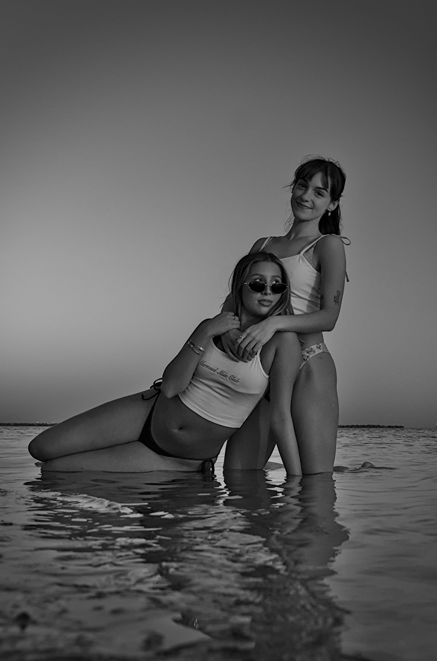 Two women pose together in shallow ocean water in a black and white beach photo wearing swimsuits.