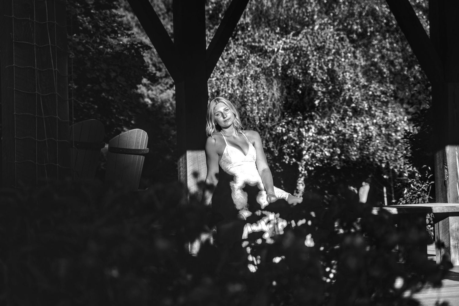 Black and white photo of a woman standing among trees and lush garden foliage in soft sunlight.