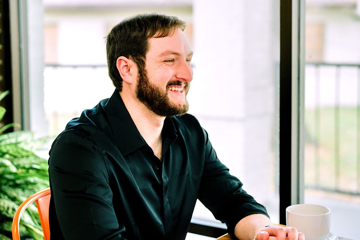 Smiling bearded man in black shirt sitting at a table near bright windows in a modern cafe setting.