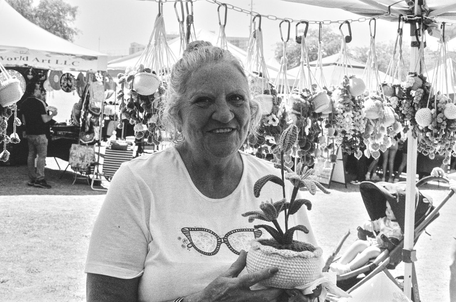 Elderly woman smiling at outdoor craft fair holding crocheted plant pot, black and white photo.