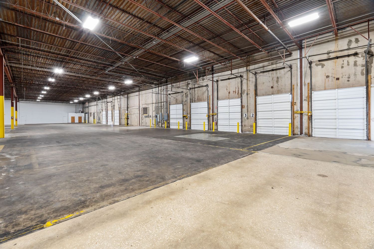 Large empty industrial warehouse interior with concrete floors and exposed ceiling beams illuminated by bright lights.