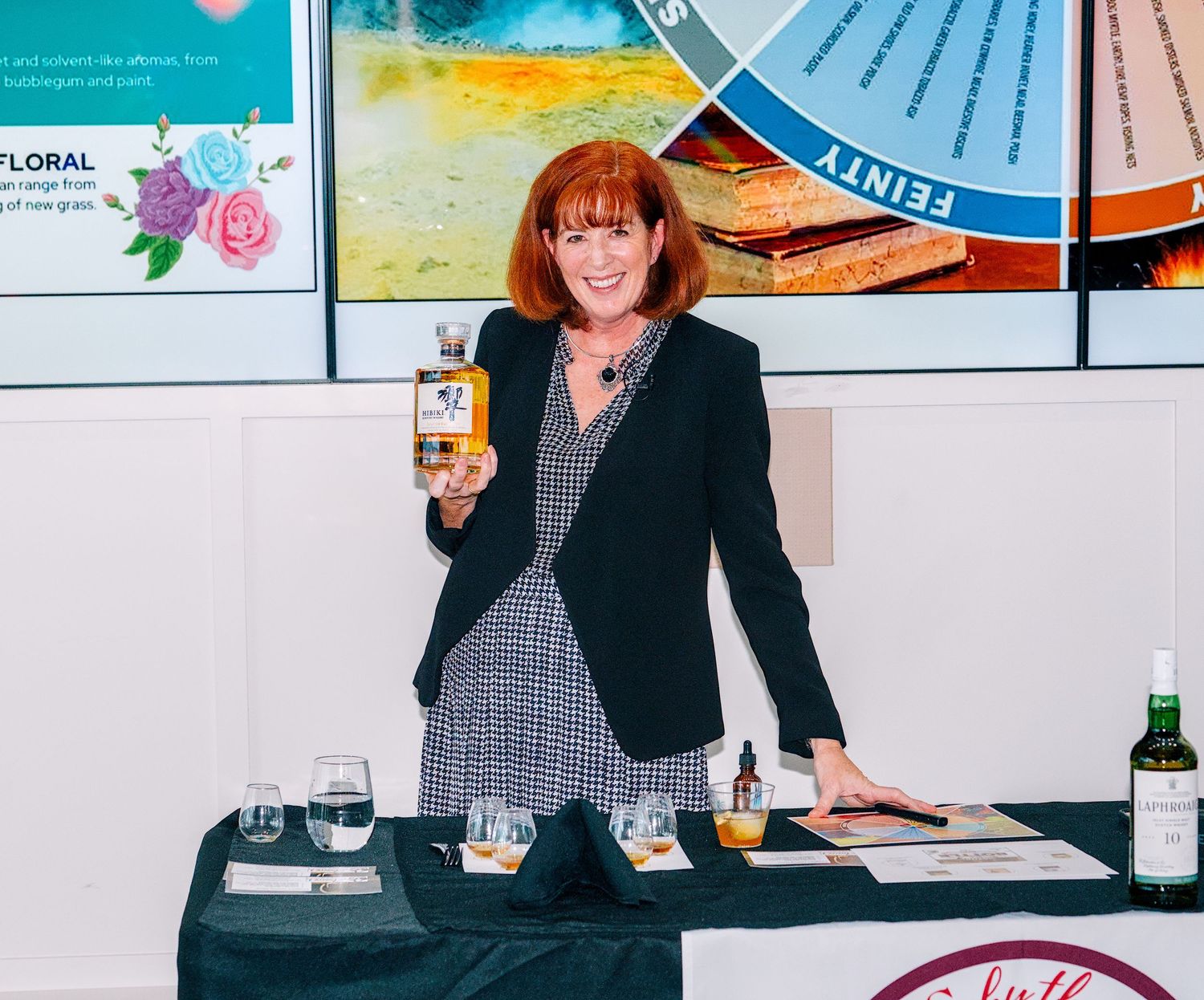 Woman presenting whisky bottle at tasting event with flavor wheel display and tasting glasses on table.