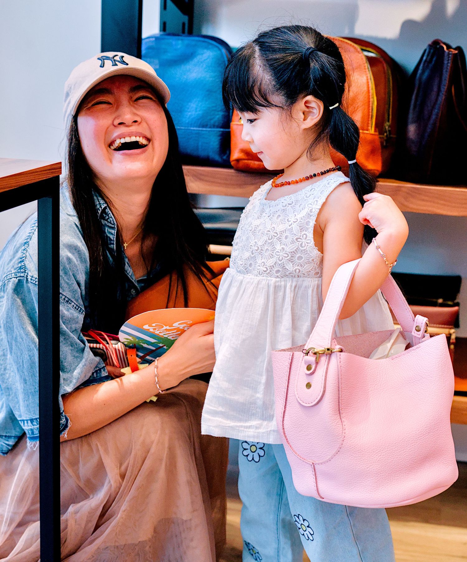 A laughing woman in an NY cap sits beside a little girl holding a pink handbag in a bag store.