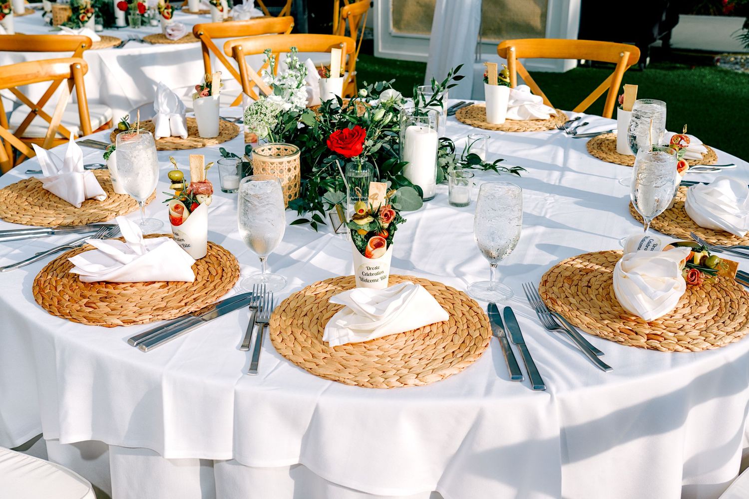 Elegant outdoor wedding reception table set with wicker placemats, white linens, and floral centerpiece.