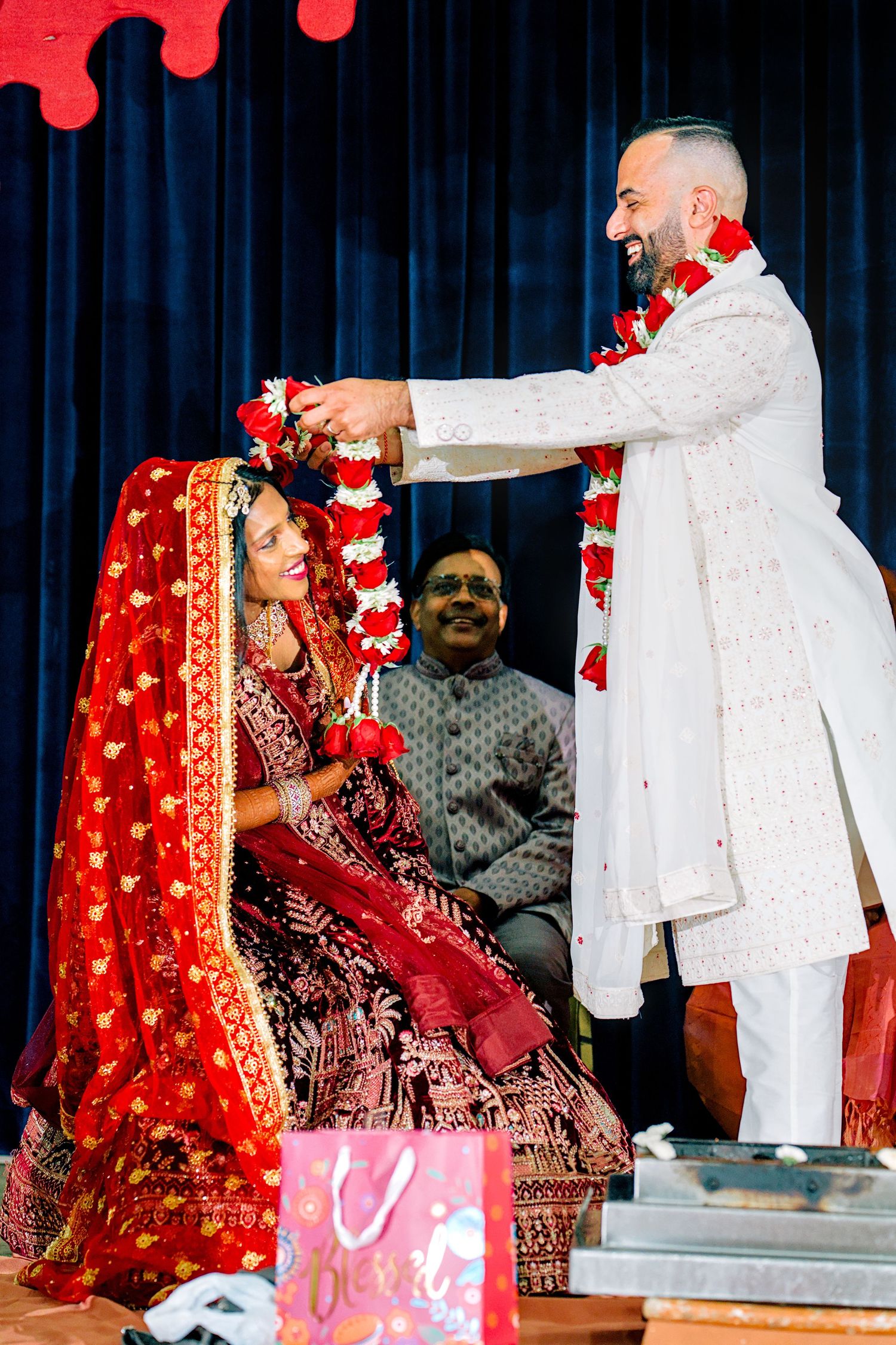 Groom in white sherwani placing floral garland on bride in red lehenga during traditional Indian wedding ceremony.