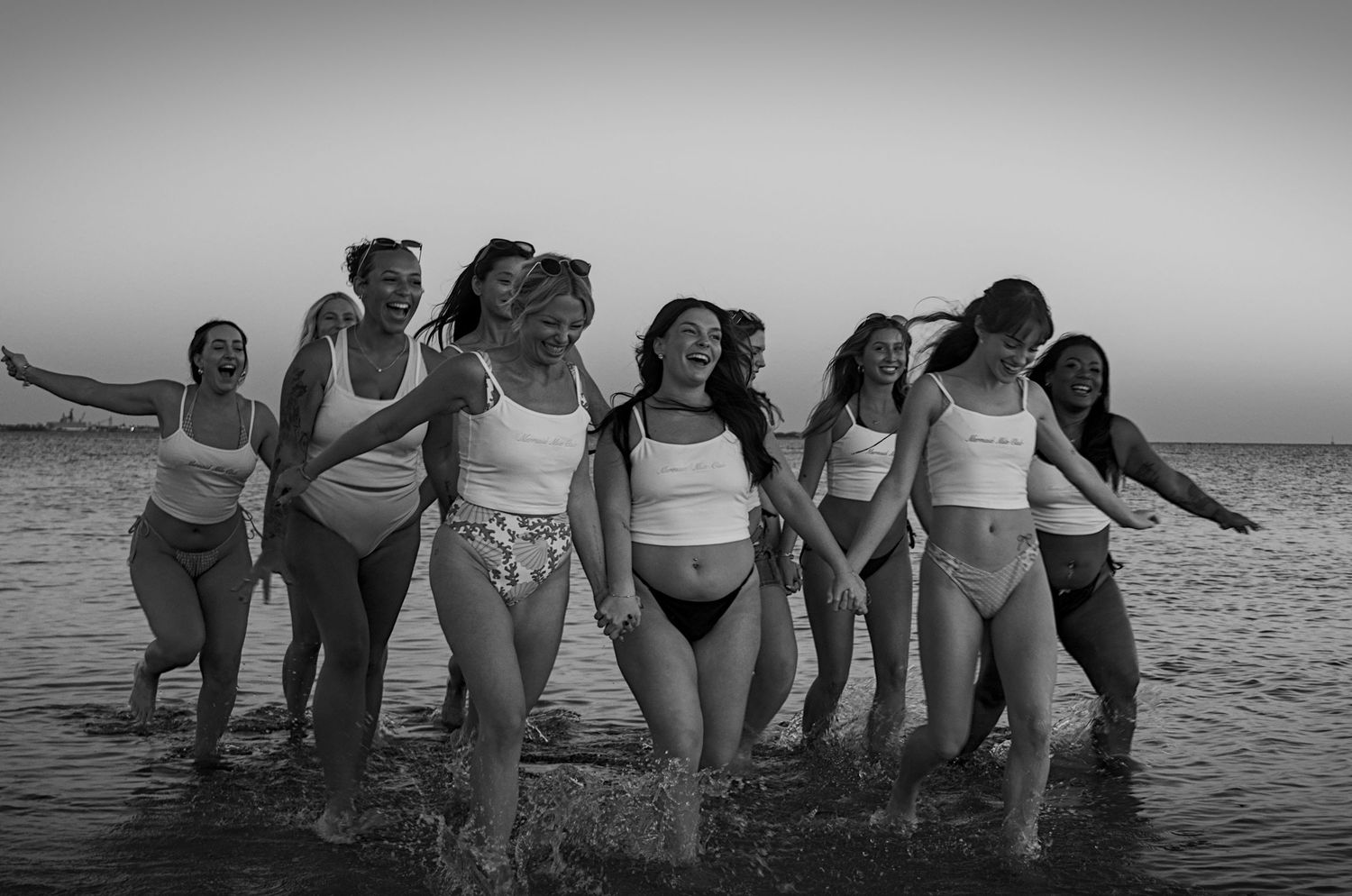 A group of women in swimwear joyfully splashing together in the ocean water at sunset in black and white photography.
