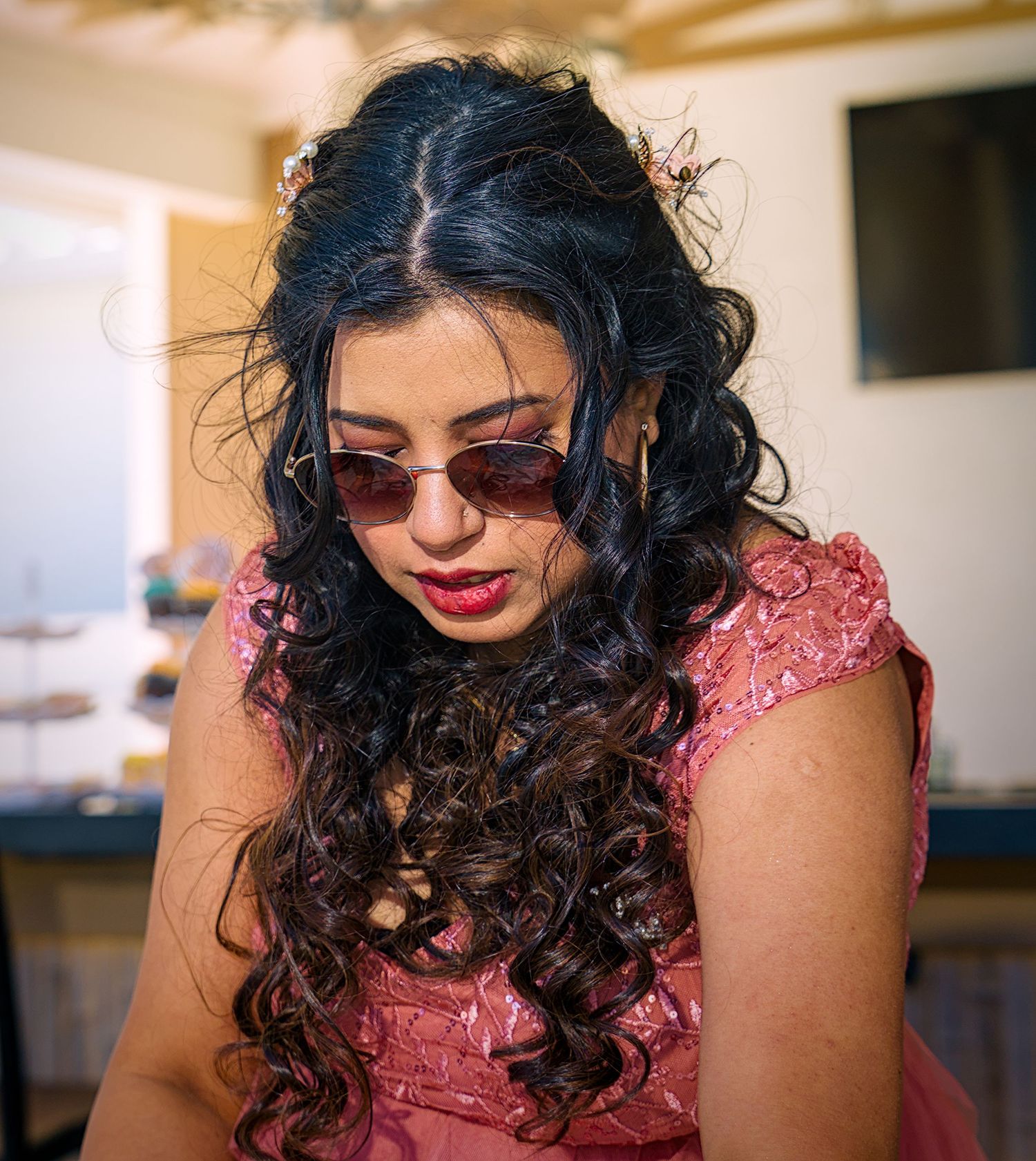 A glamorous look featuring dark curly hair, sunglasses and a pink sequined dress in an indoor setting.