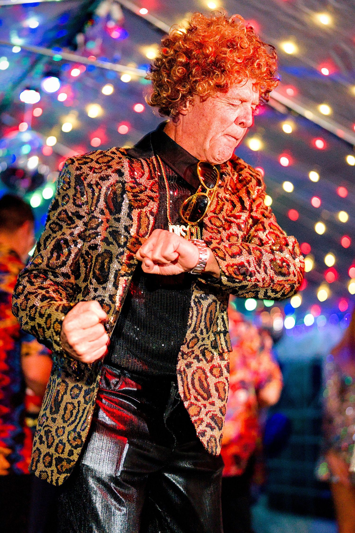 Person in leopard print jacket and red curly wig dancing under festive lights.