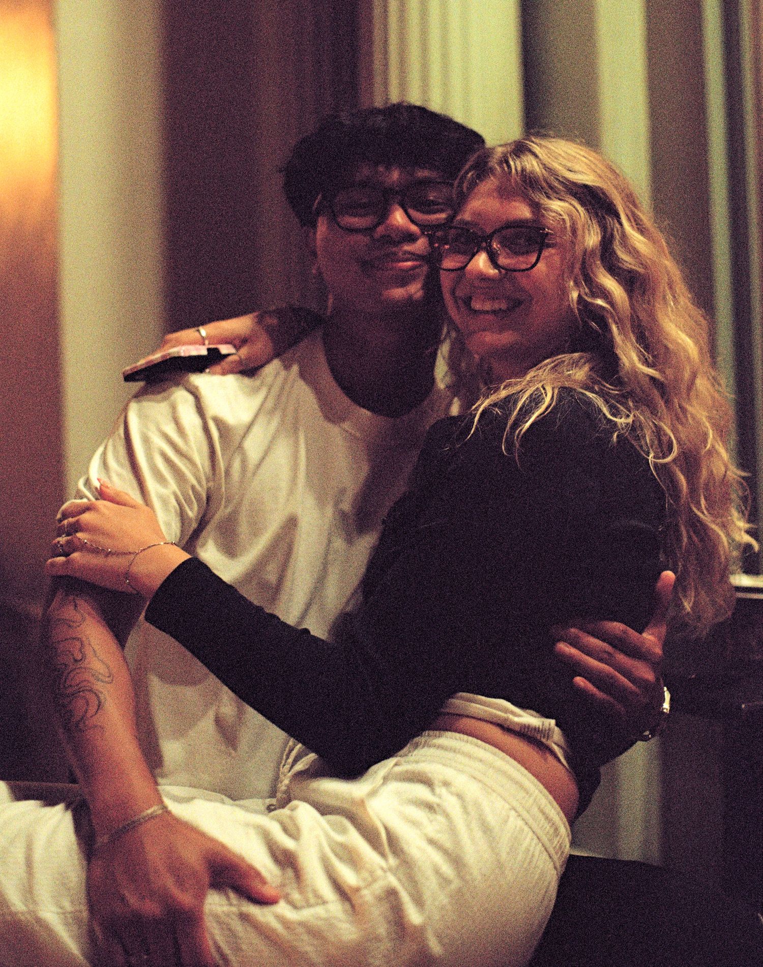 Two friends smiling and hugging, both wearing glasses, in a warmly lit indoor setting.