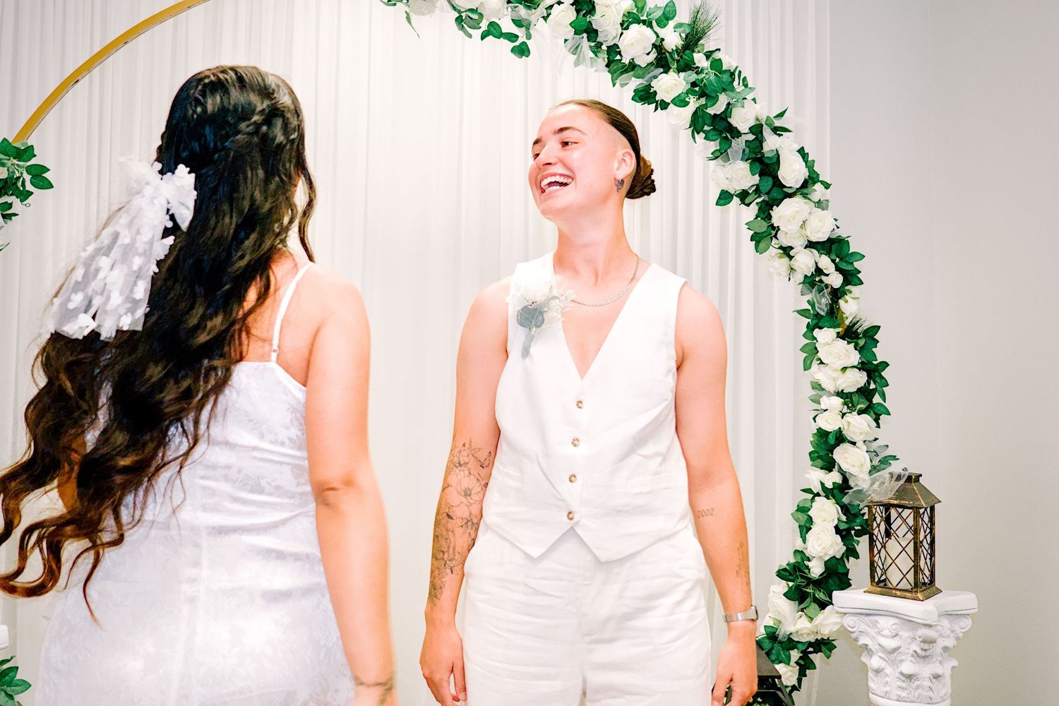 Bride in white vest laughs joyfully at altar with floral arch during same-sex wedding ceremony.