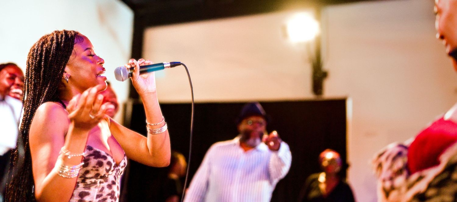 A woman singer performs passionately into a microphone at a live music event with other performers in the background.
