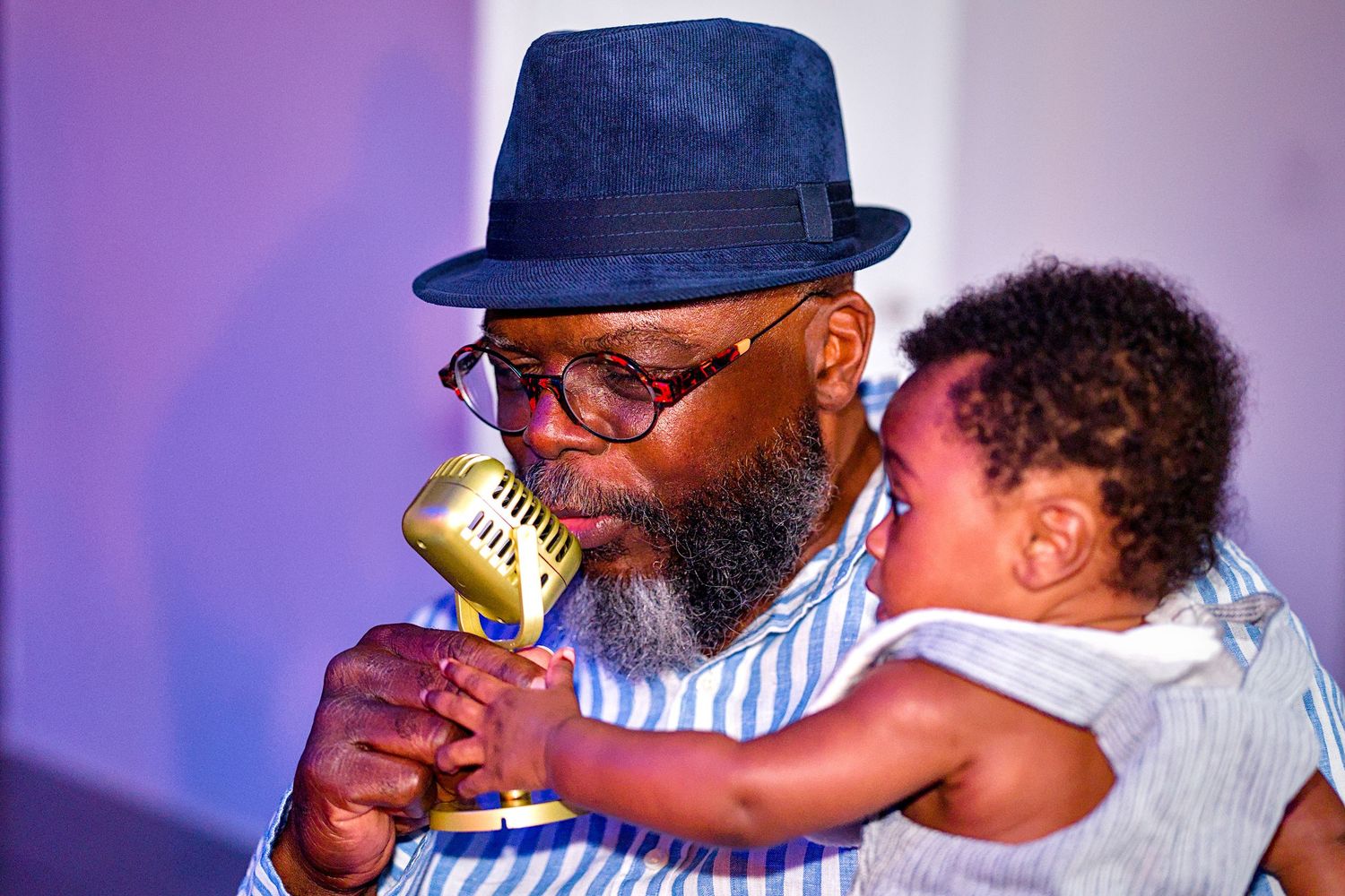 A bearded man in a blue hat holds a laughing baby while singing into a gold microphone on stage.