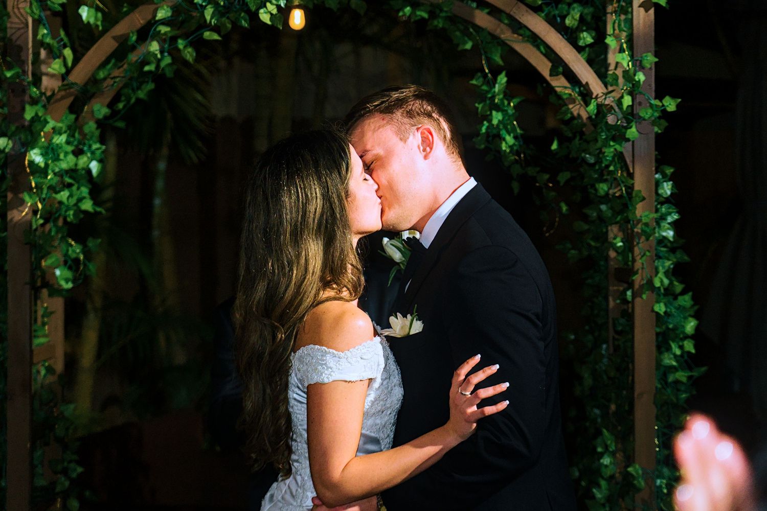 Bride and groom share first kiss under floral arch at wedding ceremony.