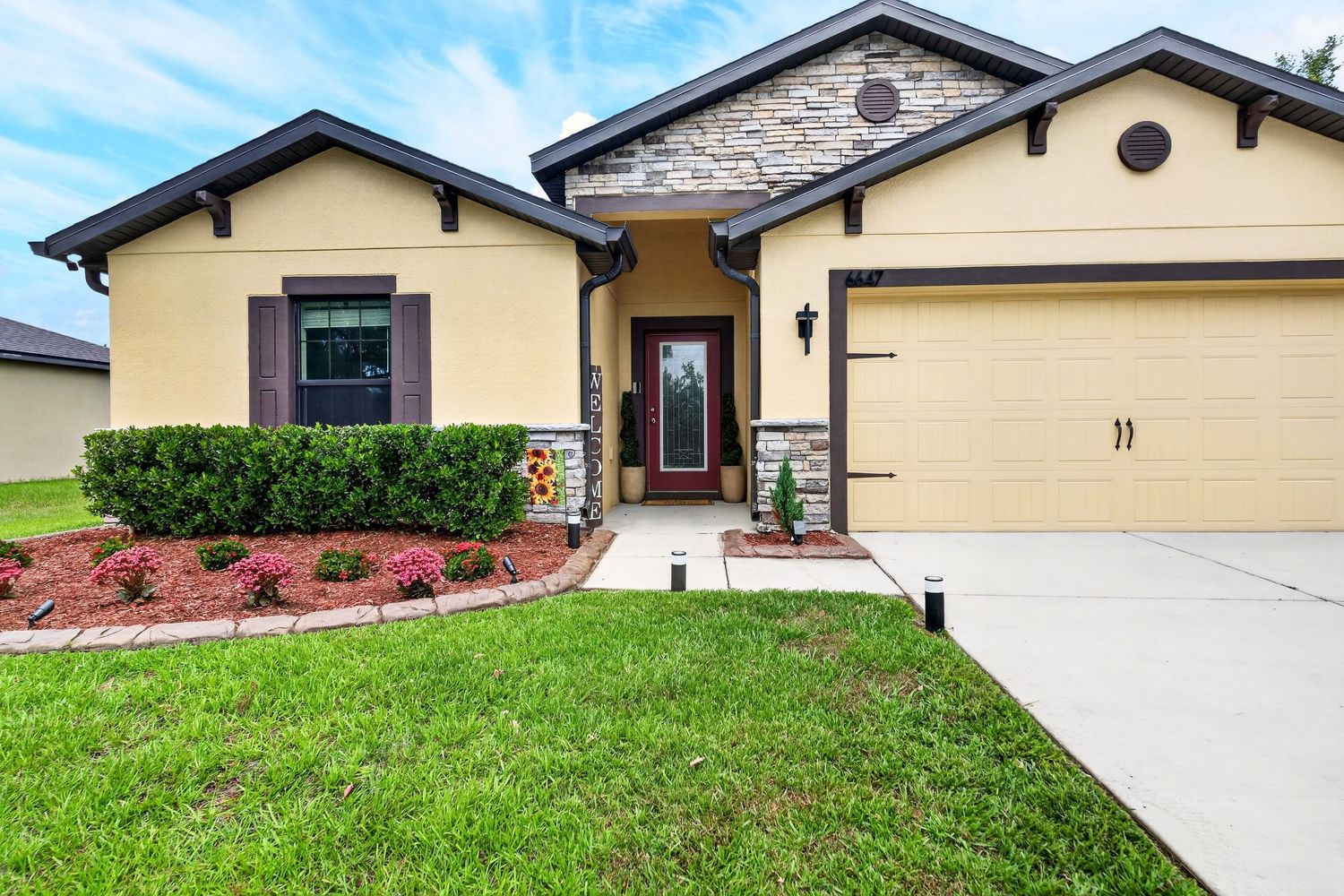 Single-story Florida home with yellow stucco exterior, stone accents, two-car garage, red front door, and lush green landscaping.