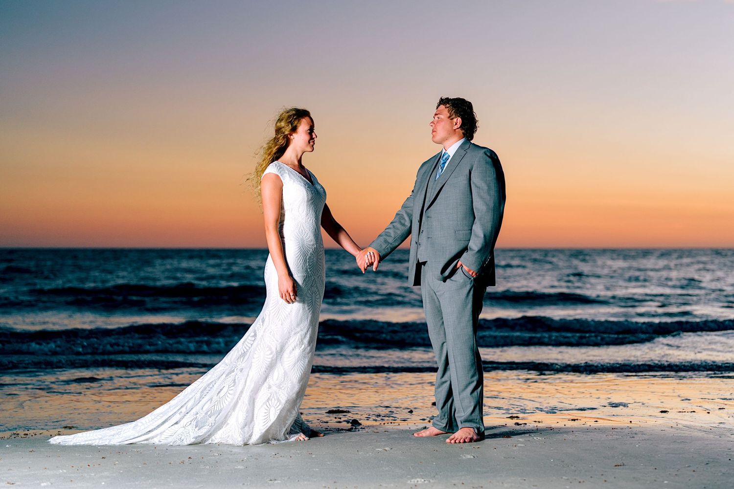 Bride and groom holding hands barefoot on a beach at sunset in elegant white dress and grey suit.