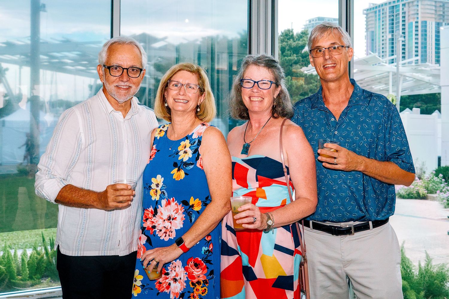 Four adults smiling and holding drinks at an indoor social gathering with large windows and city views in the background.