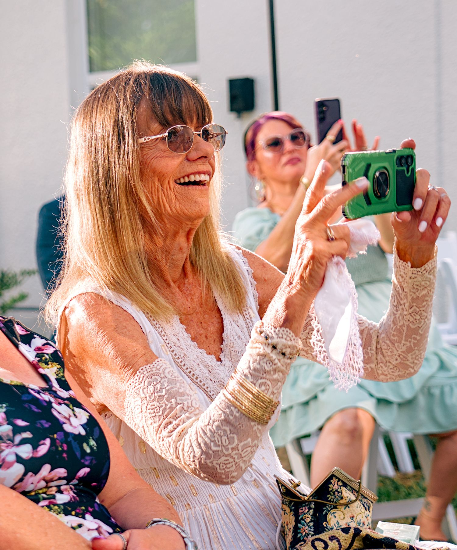 Smiling blonde woman in white lace top takes selfie on green phone at outdoor wedding reception.