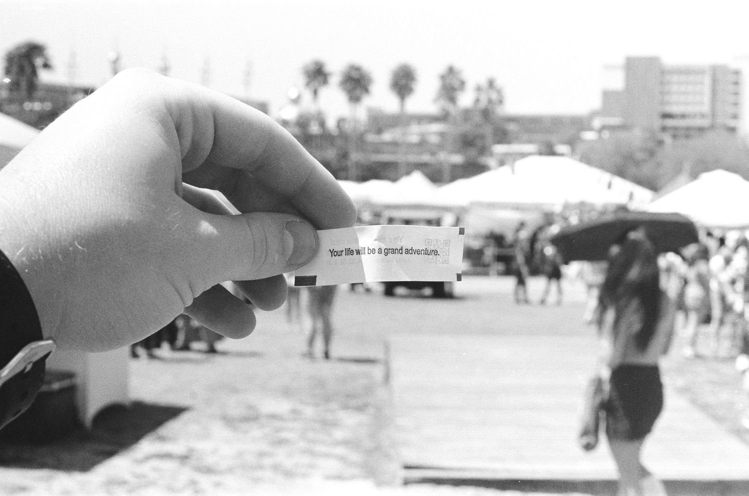 Person holding a small ticket or wristband at an outdoor event with palm trees and crowd in black and white photo.