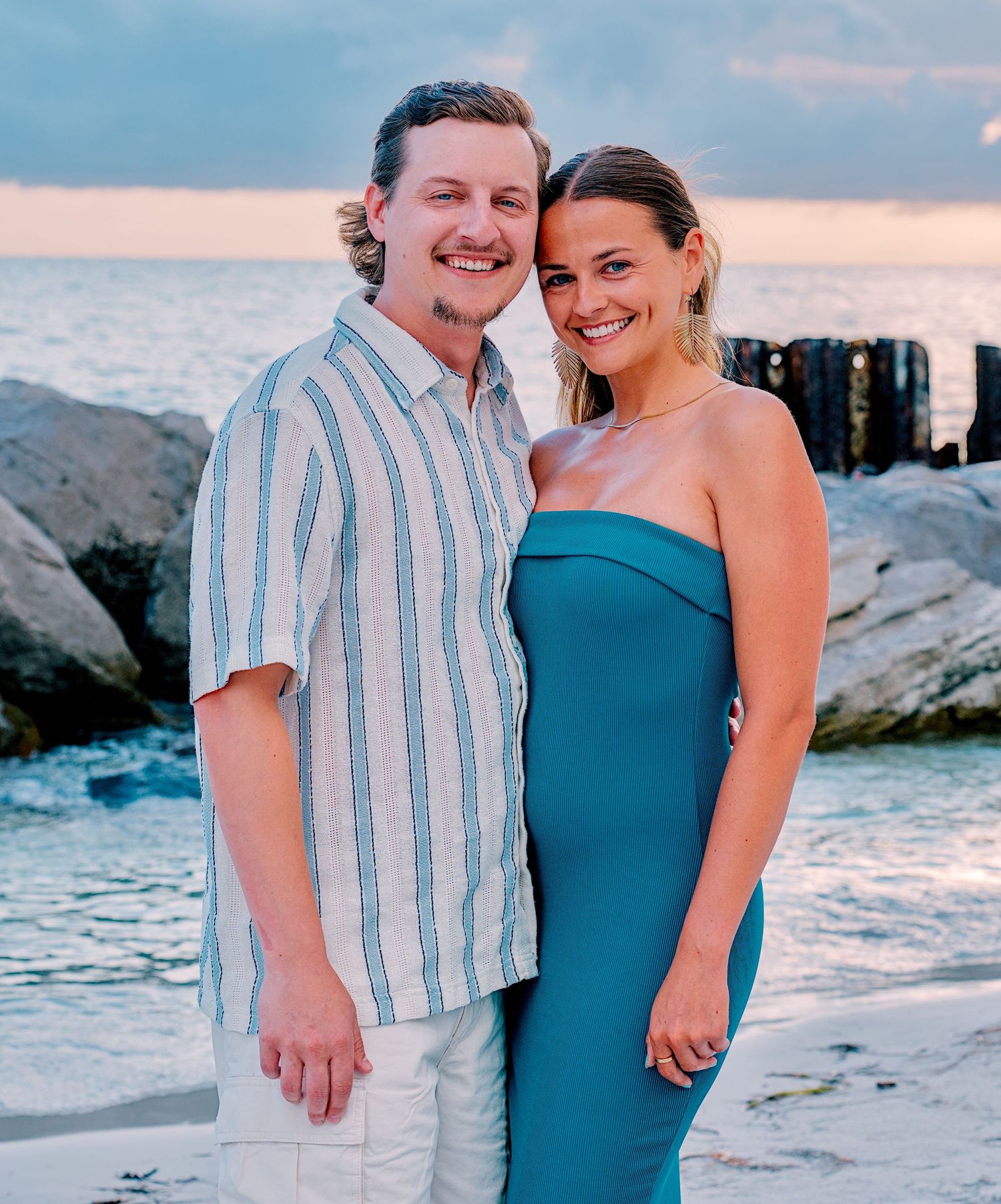 Couple smiling together on a rocky beach at sunset, man in striped shirt and woman in teal strapless dress.