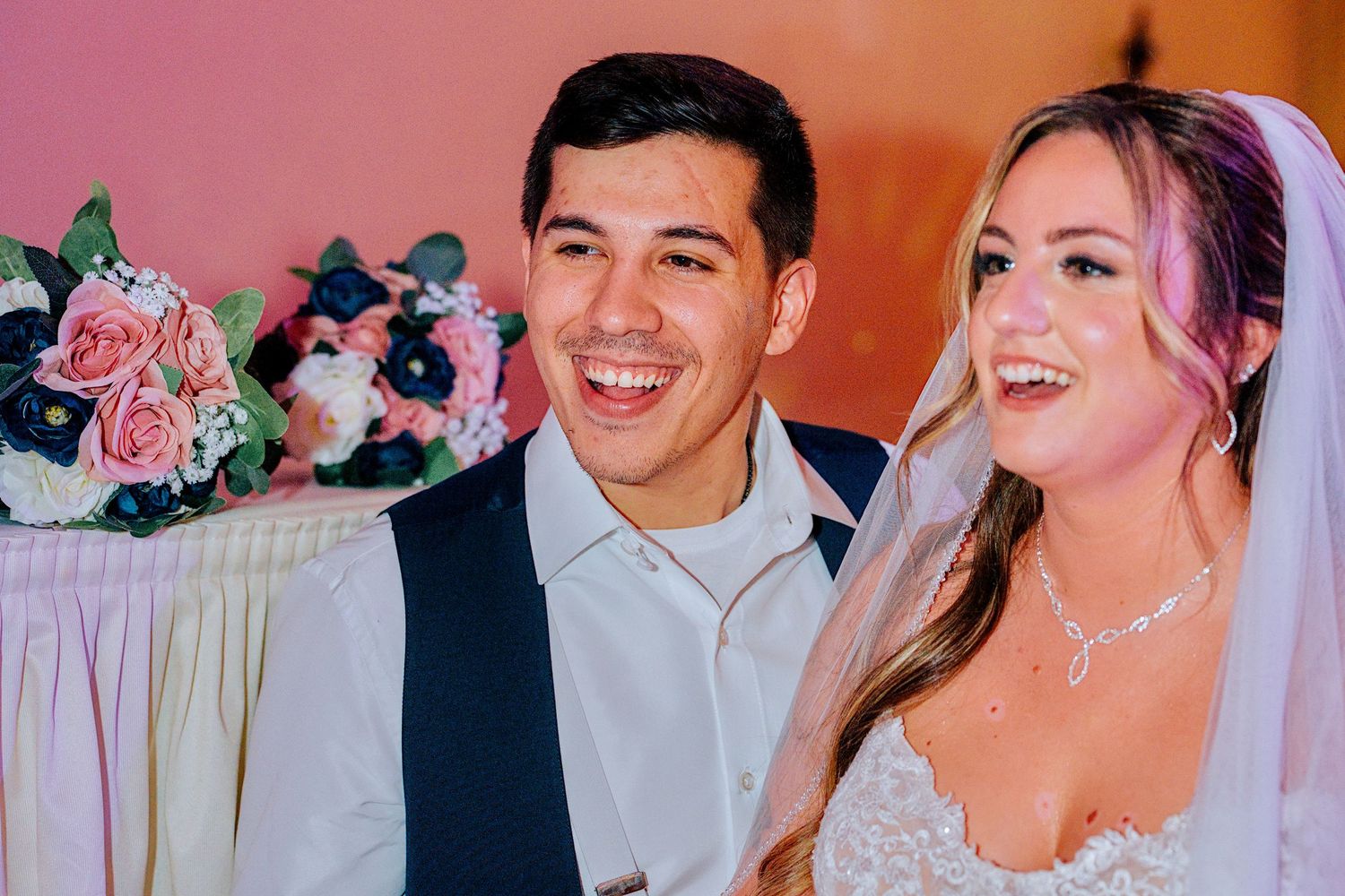 Smiling bride and groom sit together at wedding reception with floral arrangement behind them.