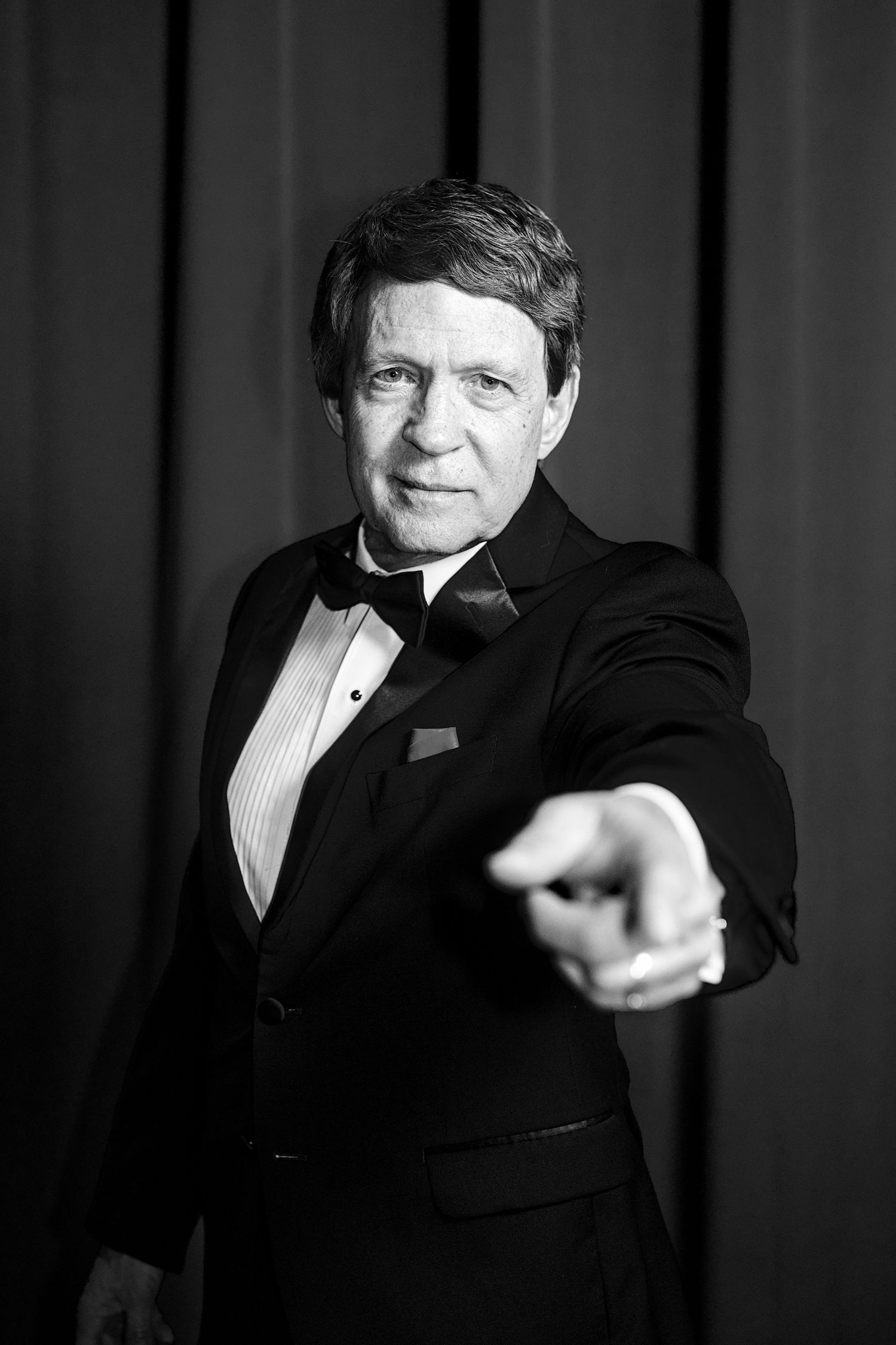 Black and white portrait of an older man in a tuxedo pointing at the camera against a dark curtain backdrop.
