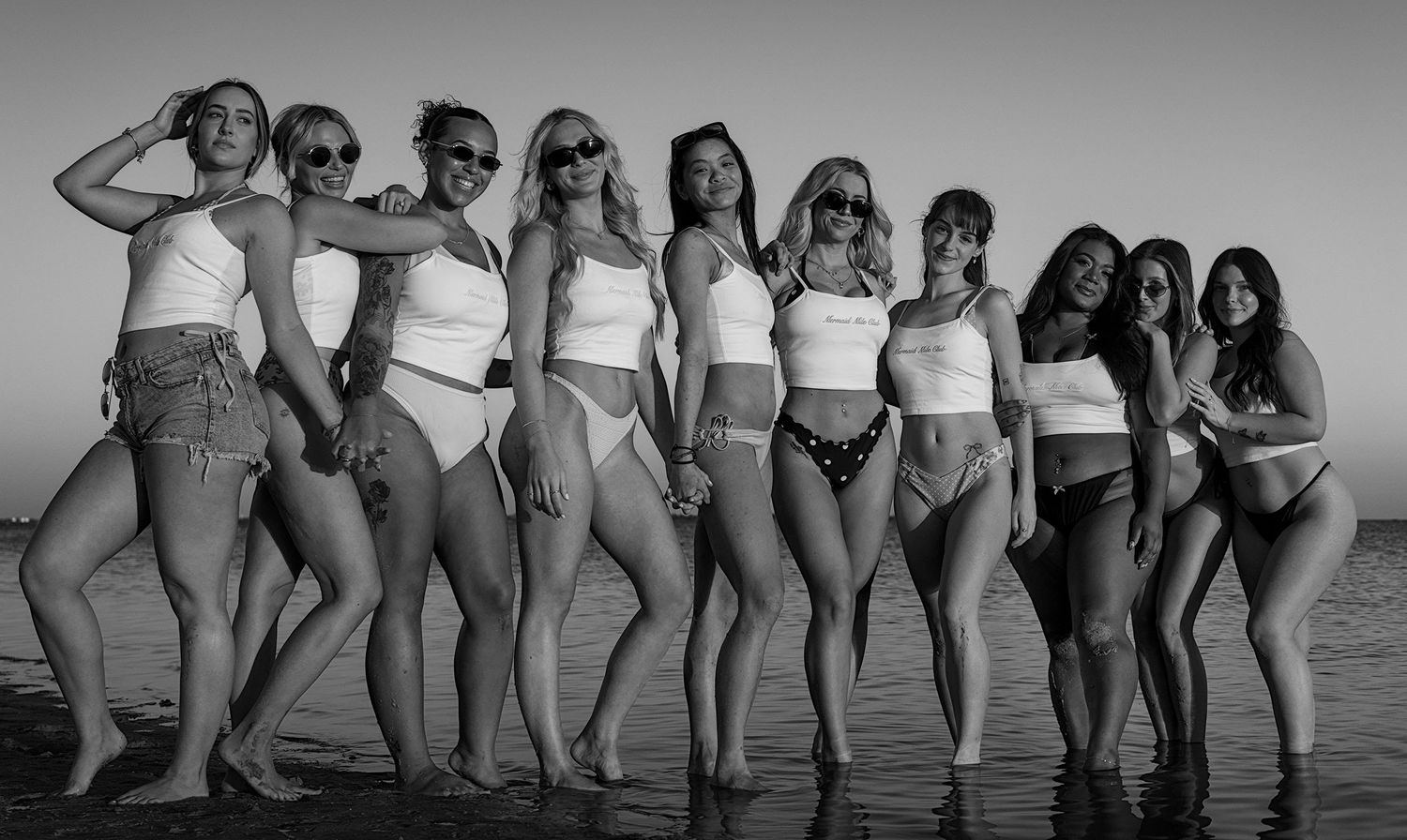 Black and white photo of a group of women posing together on a beach in swimwear and casual summer outfits.