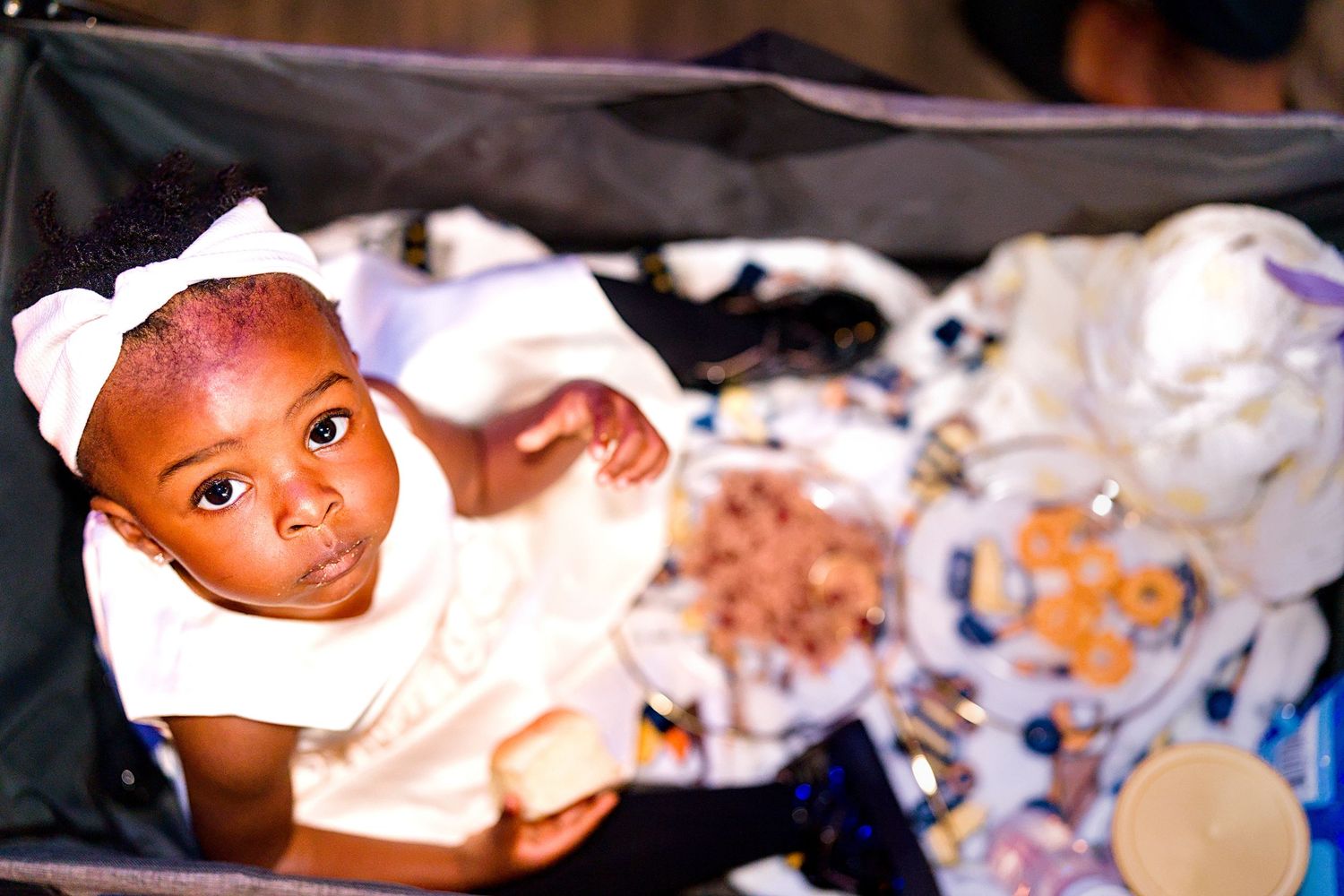 Cute Black baby in a white bow lies in a stroller surrounded by colorful blankets and items.