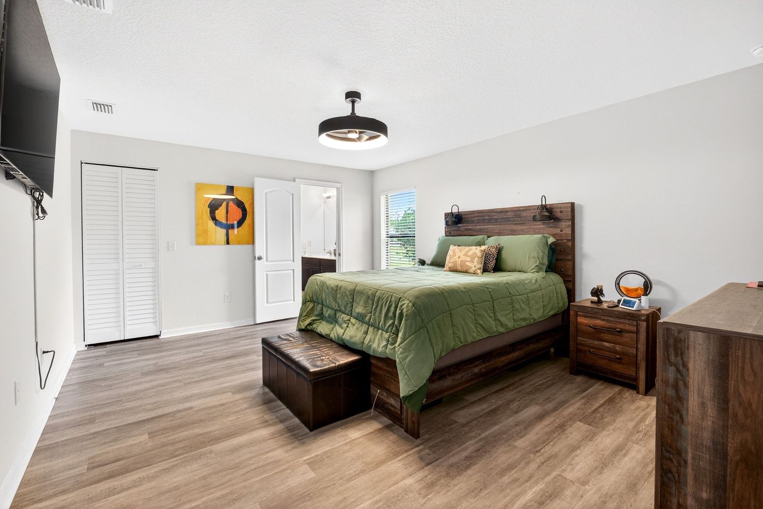 Spacious bedroom with hardwood floors, dark wood bed frame, green bedding, white closet doors, and modern ceiling fan.