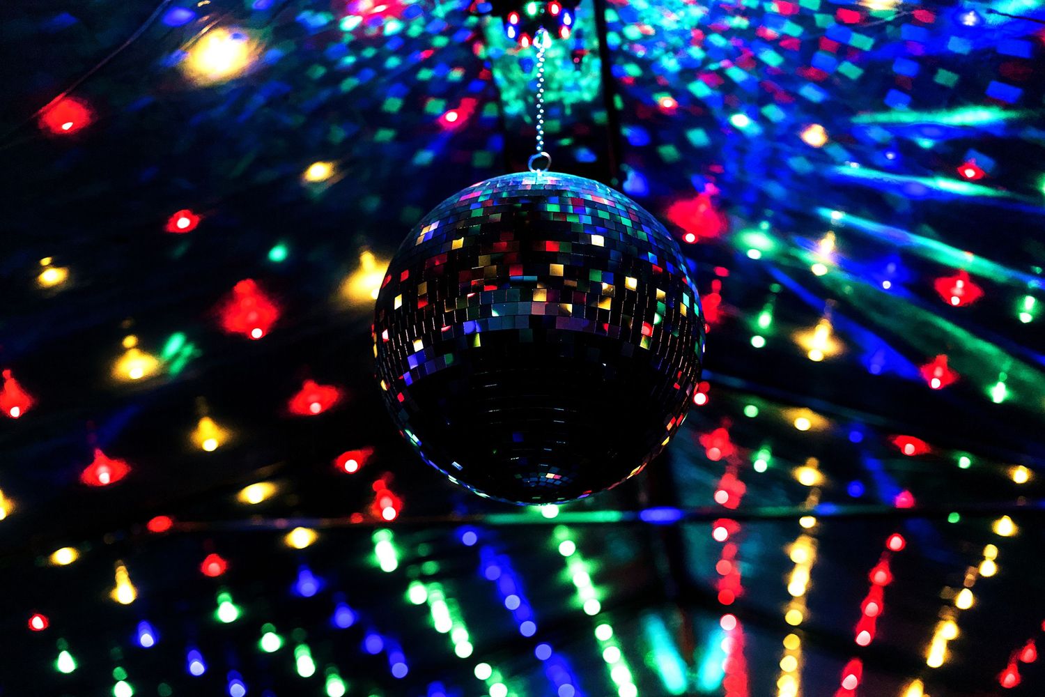 A shimmering disco mirror ball hanging from the ceiling surrounded by colorful bokeh lights in a dark party venue.