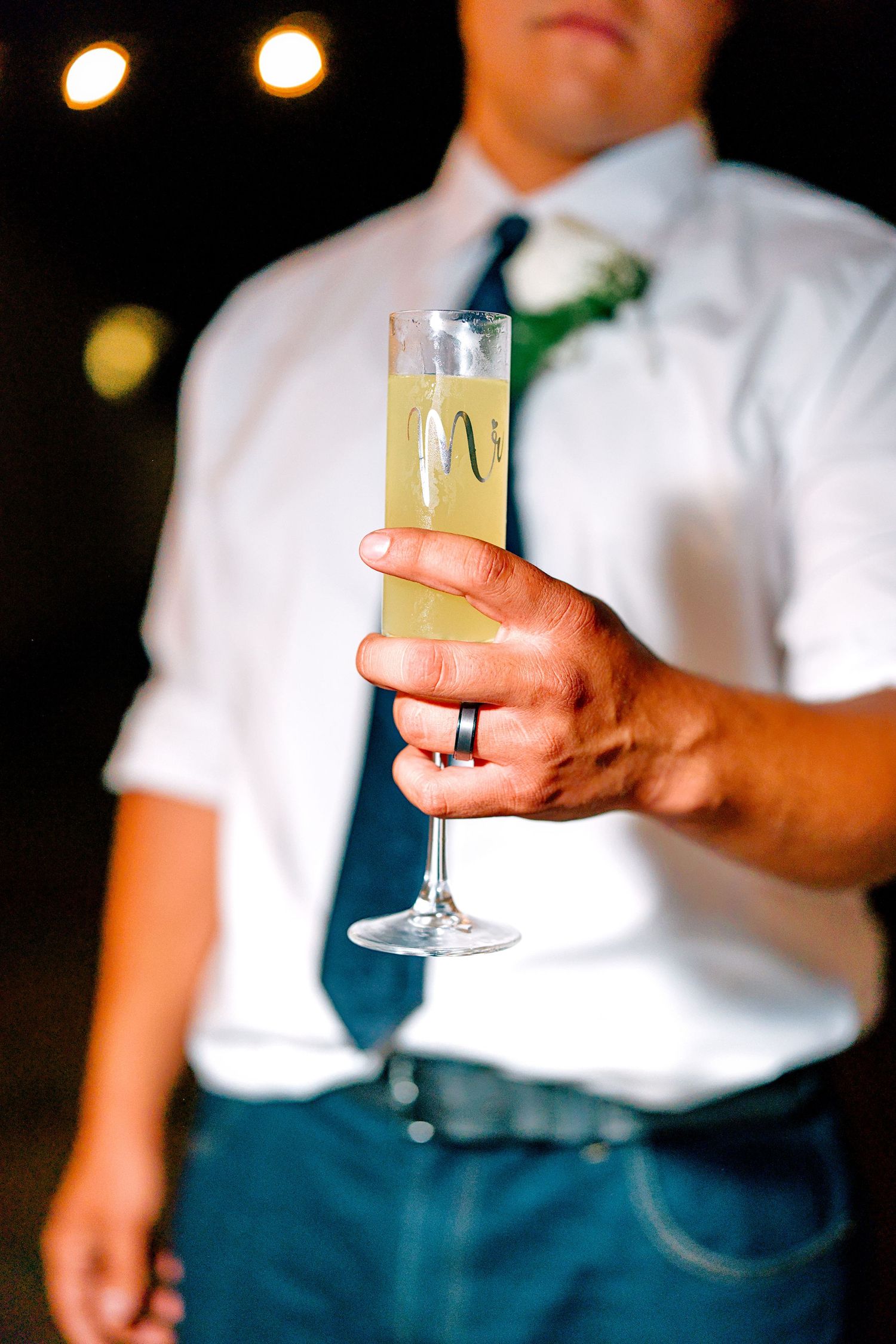 A groom in a white shirt and blue tie holds a monogrammed champagne flute at his wedding reception.