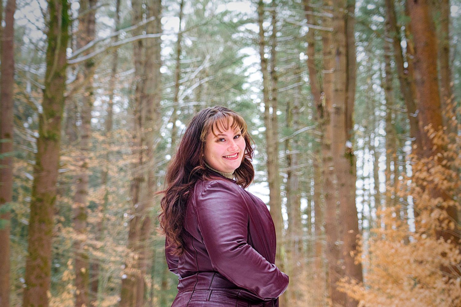 A person in a burgundy jacket poses against autumn trees in a forest setting.