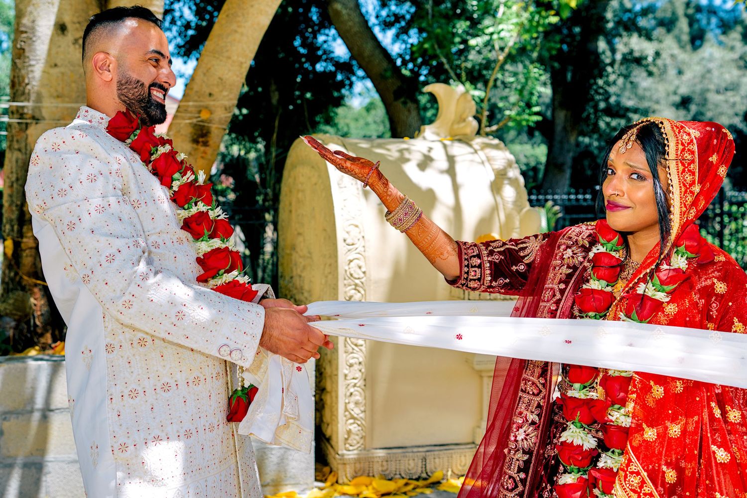 Indian bride in red sari and groom in white sherwani exchange garlands at traditional Hindu wedding ceremony outdoors.